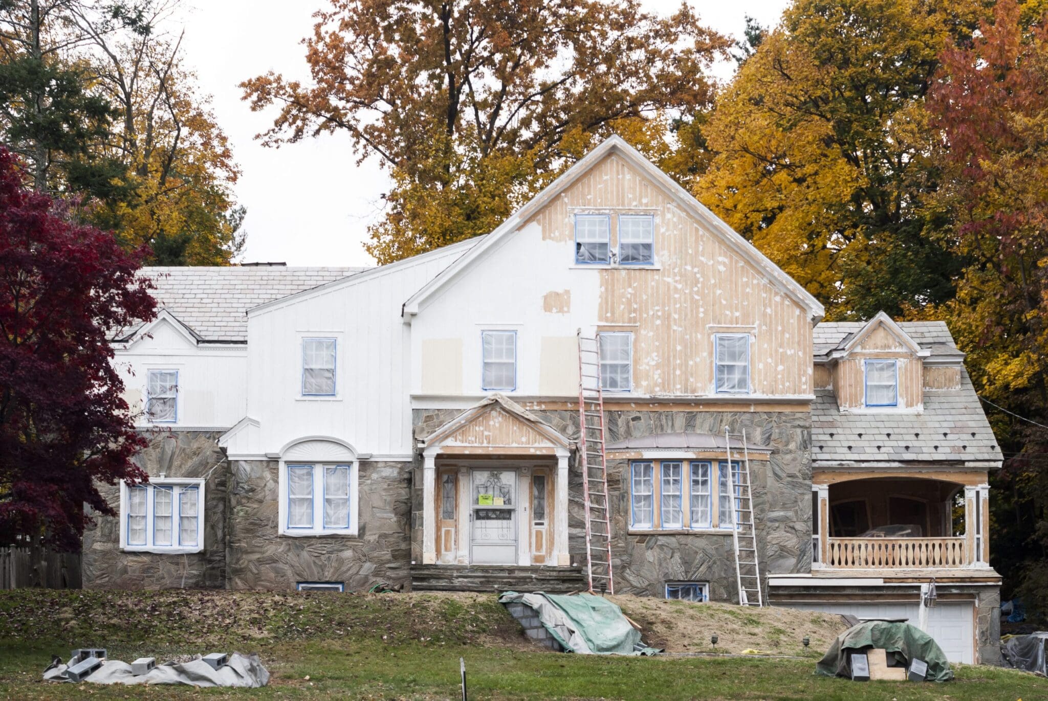 A home has ladders leaning against it, with half of the house having a fresh coat of white paint applied to it, while the other half is a faded brown color, waiting to be repainted.